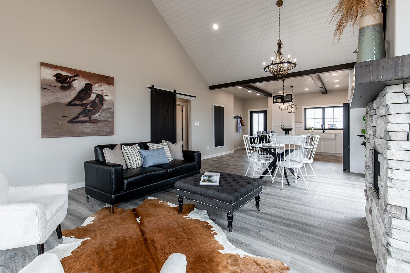 Spacious living area with high ceilings, featuring a black leather sofa, white chairs, cowhide rug, and stone fireplace. Rustic chandelier and beam accents.