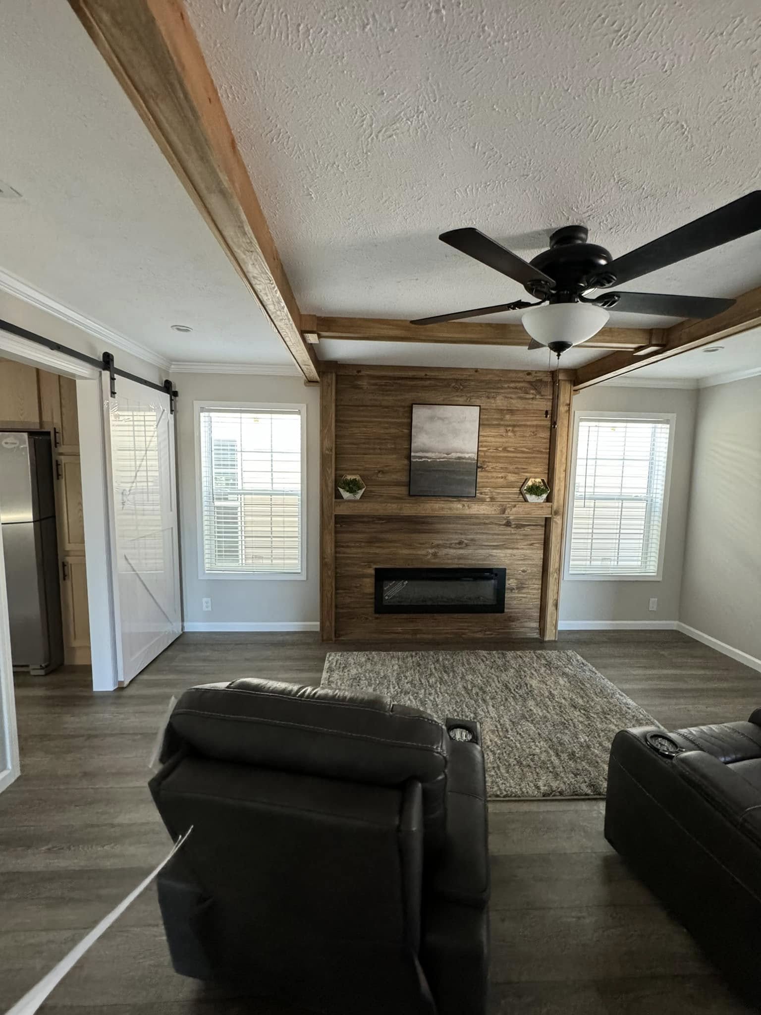 Cozy living room with a wooden accent wall featuring a modern fireplace. Two recliners face a rug; ceiling fan above and barn door beside windows.