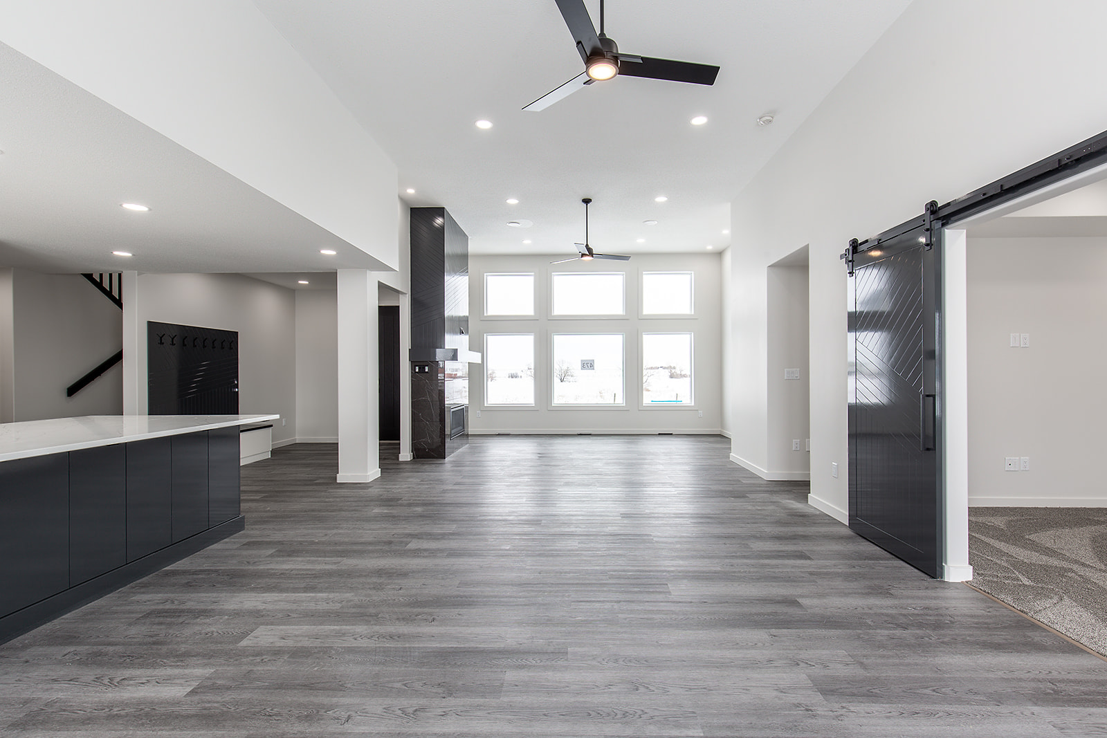 Spacious, modern interior with gray wooden flooring, white walls, large windows, ceiling fans, kitchen area with black cabinets, and sliding barn door.