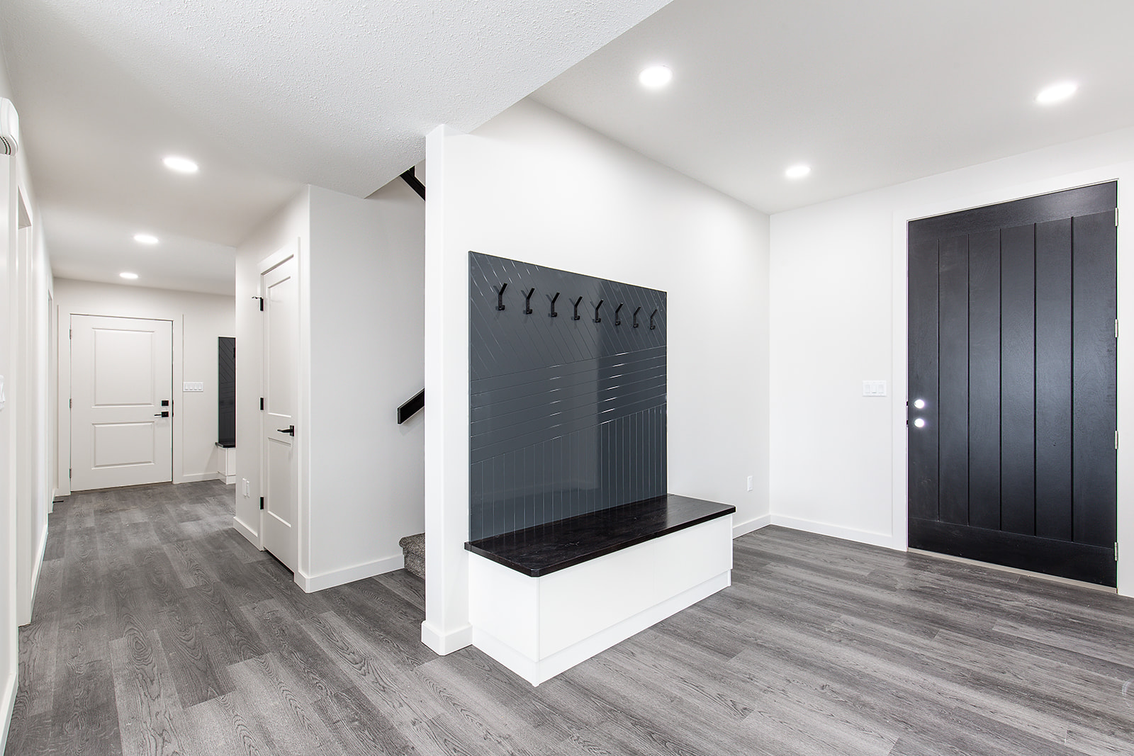 Modern entryway with gray wood flooring, white walls, and recessed lighting. Features a black bench with coat hooks and a dark wooden door. Minimalist design.