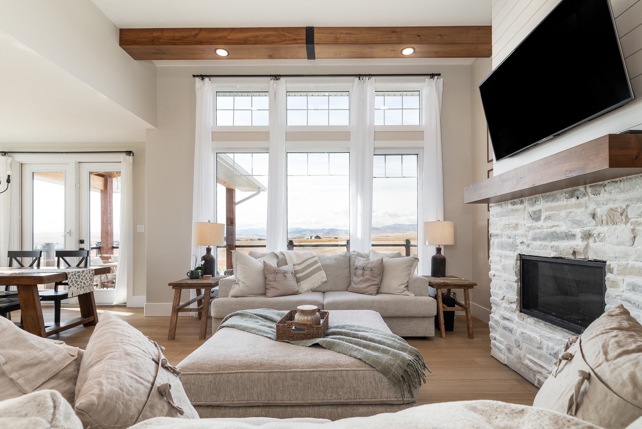 Bright, cozy living room with large windows overlooking mountains. Features neutral decor, stone fireplace, cushioned sofa, and wooden beams. Warm, inviting ambiance.