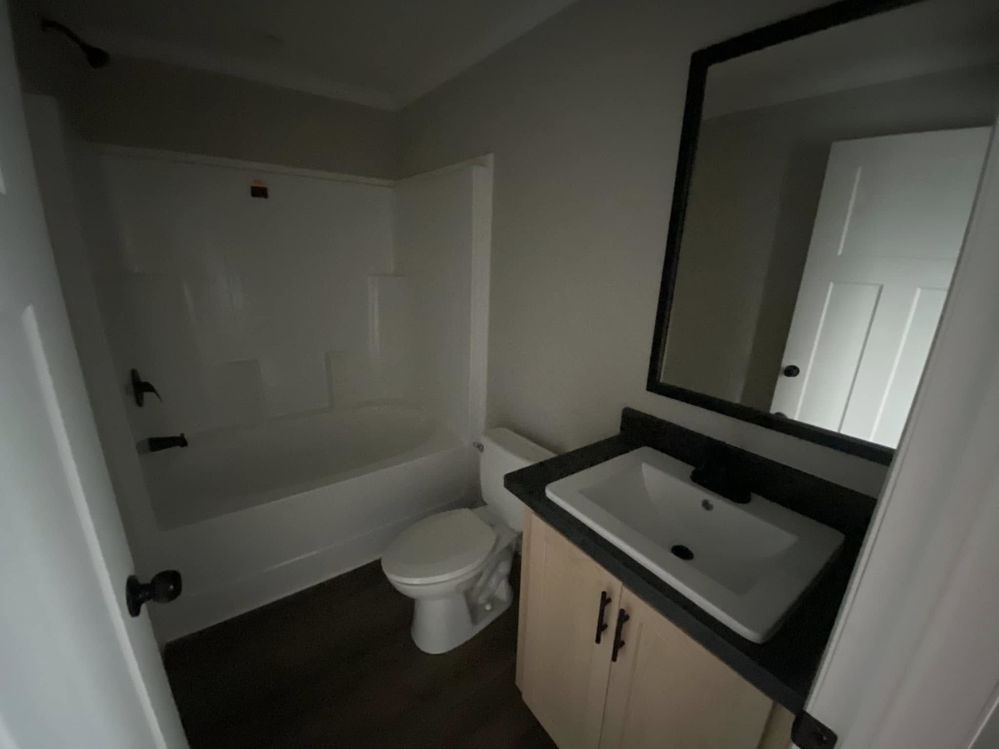 Compact bathroom with a white bathtub, toilet, and a wooden vanity featuring a black countertop and sink. A large mirror hangs above the vanity.