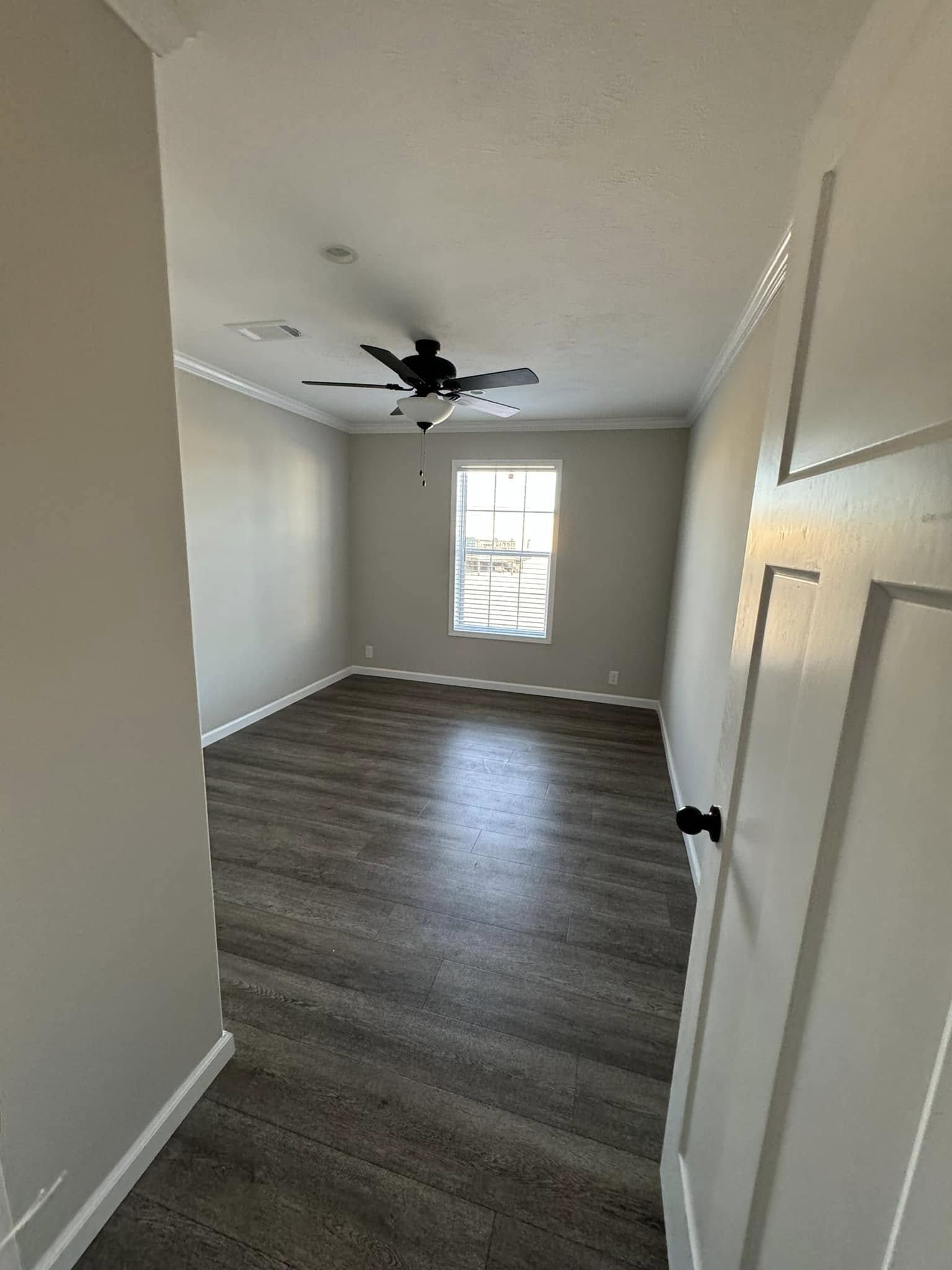 Empty room with light gray walls and dark wood flooring, featuring a central window and ceiling fan. The space feels bright and minimalist.