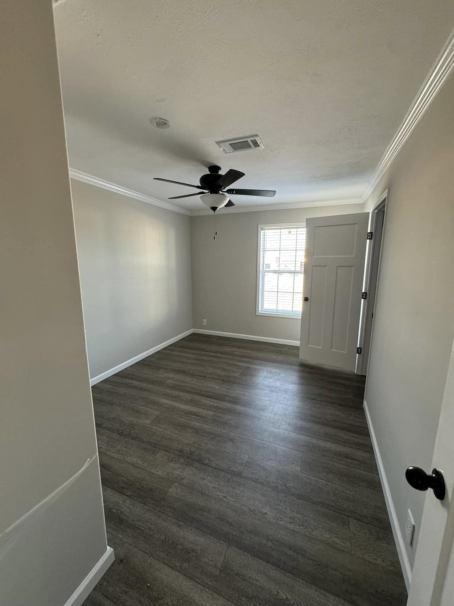 An empty room with gray walls and dark wood vinyl flooring. A ceiling fan with light is centered overhead. Sunlight streams through a window with white blinds, creating a calm, neutral tone.