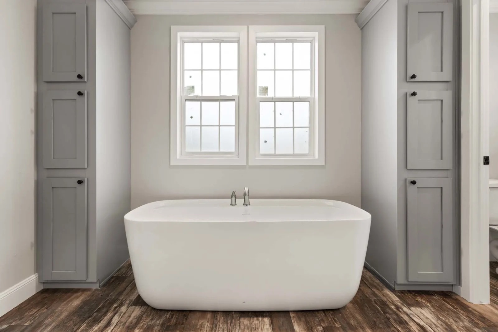 Modern bathroom featuring a freestanding white bathtub, centered below two tall windows. Flanked by gray cabinets, set on a wood-textured floor.