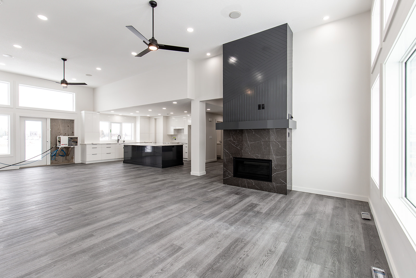 Spacious modern living room with gray wood flooring, large windows, and a sleek black fireplace. Open kitchen area features white cabinetry. Minimalist and bright.