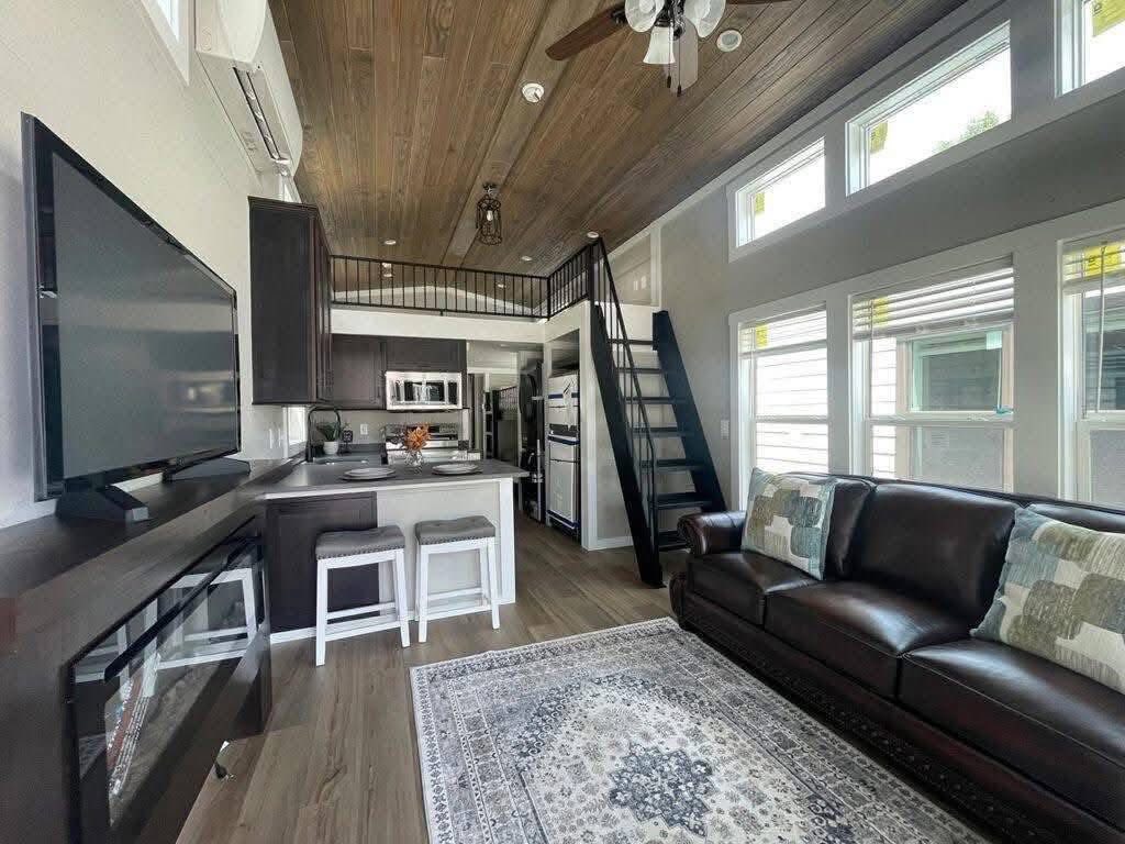Modern tiny home interior featuring a cozy living area with a dark leather sofa and patterned rug, compact kitchen with white stools, and wooden loft.