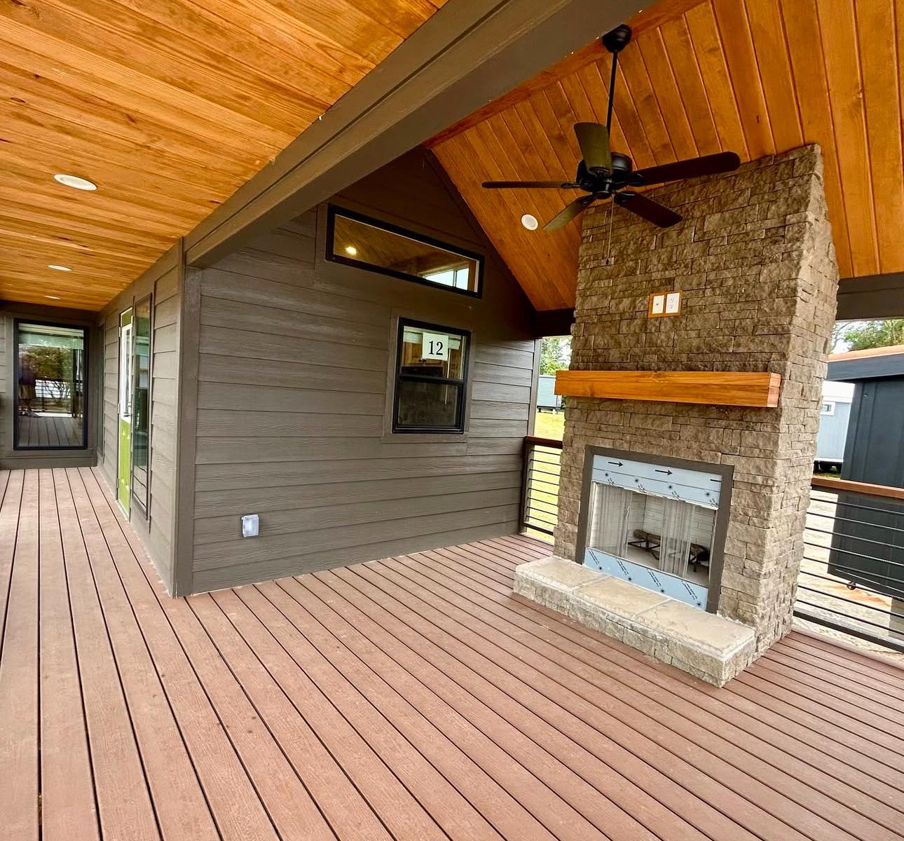 Covered patio with warm wood ceiling, brown siding, and a stone fireplace. A black ceiling fan hangs above, creating a cozy, inviting feel.