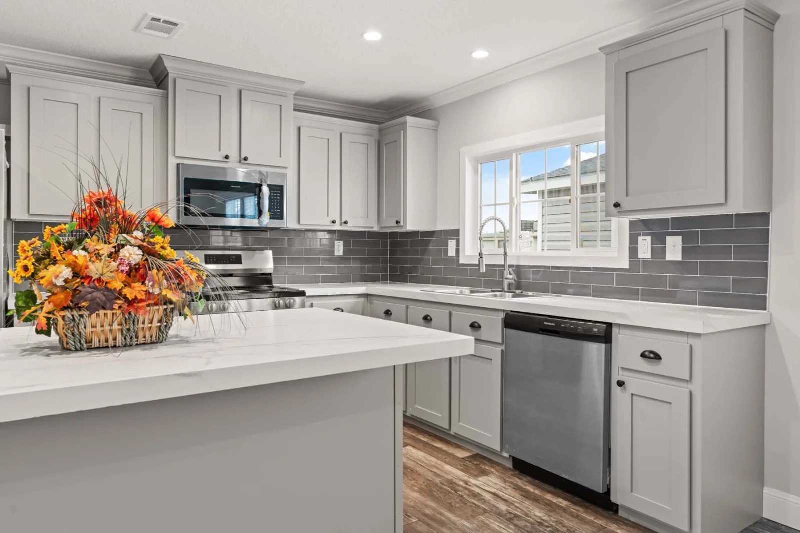 A modern kitchen with gray cabinets, white countertops, and stainless steel appliances. A basket of vibrant fall flowers adds warmth on the island.