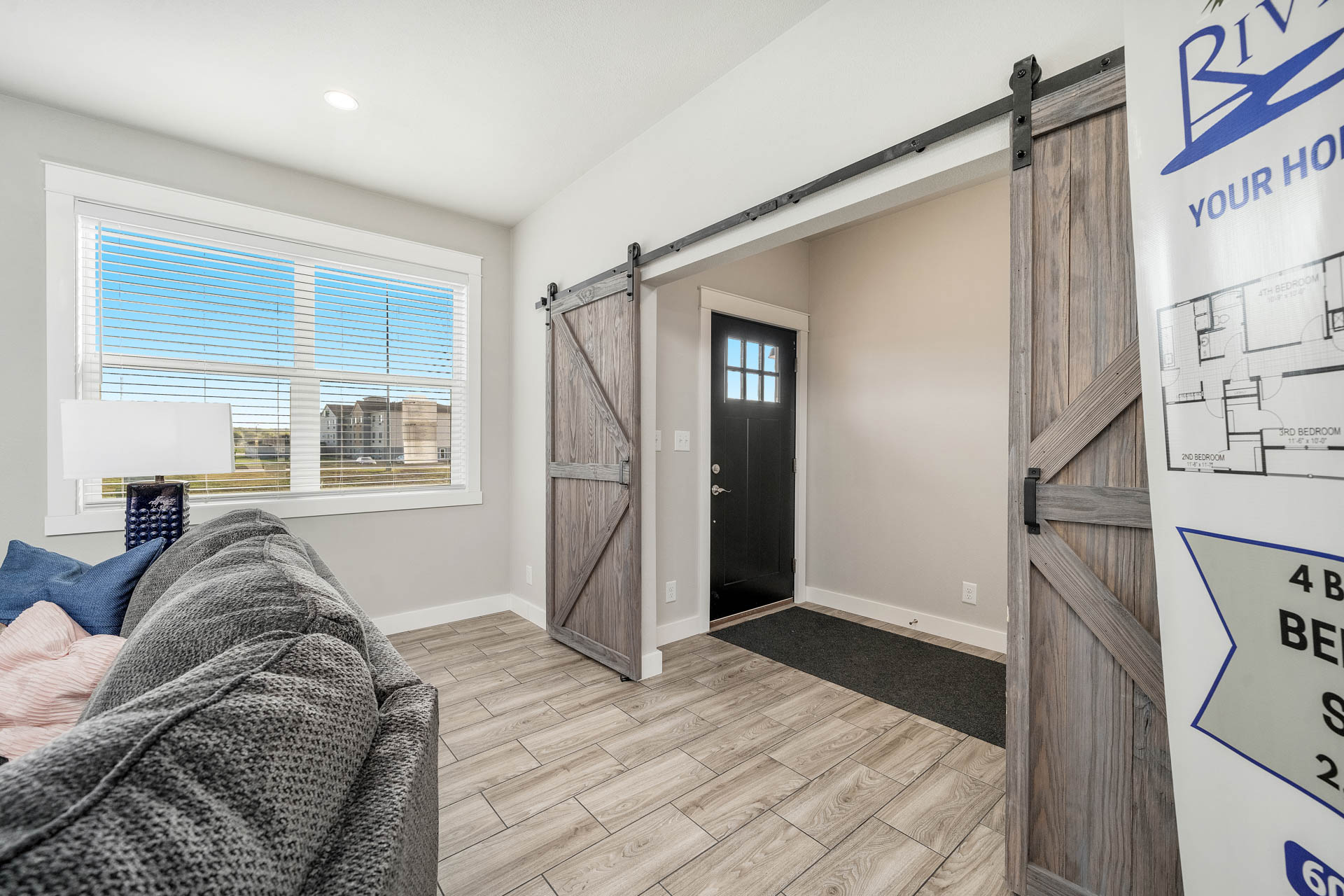 Bright living room with large window, gray sofa, and blue lamp. Barn-style sliding door opens to entryway with black door. Cozy, inviting feel.
