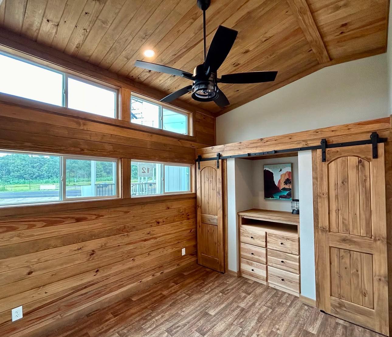 Wood-paneled room with large windows, a ceiling fan, and a rustic vibe. Features sliding barn doors revealing shelves, and artwork on the wall.