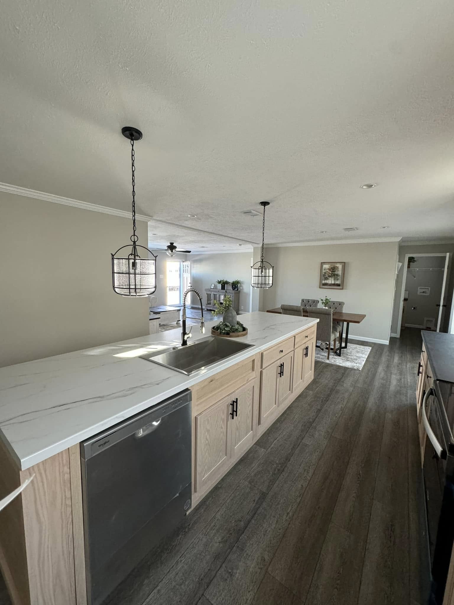Modern kitchen with a large, light wood island, marble countertop, and stainless steel sink. Pendant lights hang above. Cozy dining area in the background.