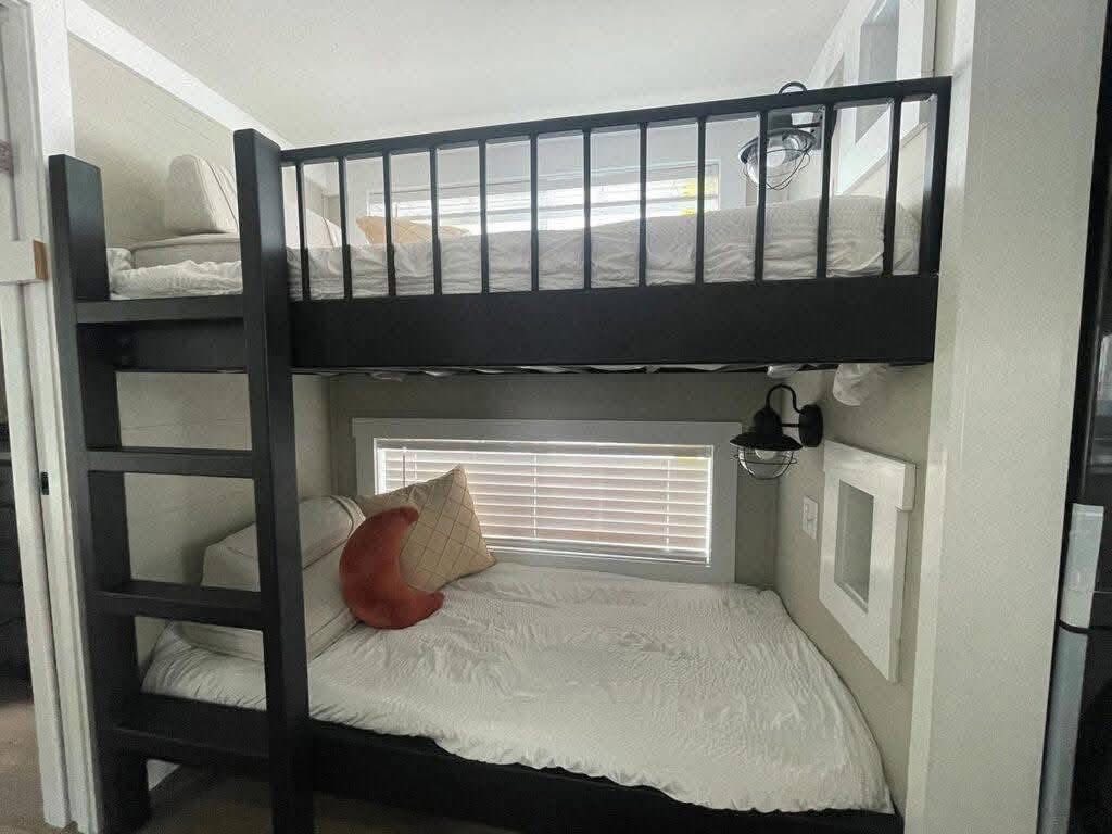 A cozy bunk bed setup in a small room, featuring black frames and white bedding. A ladder leads to the top bunk. A window with blinds is behind the lower bunk, with pillows, including a red crescent-shaped one. A wall lamp adds soft lighting, creating a warm, inviting atmosphere.