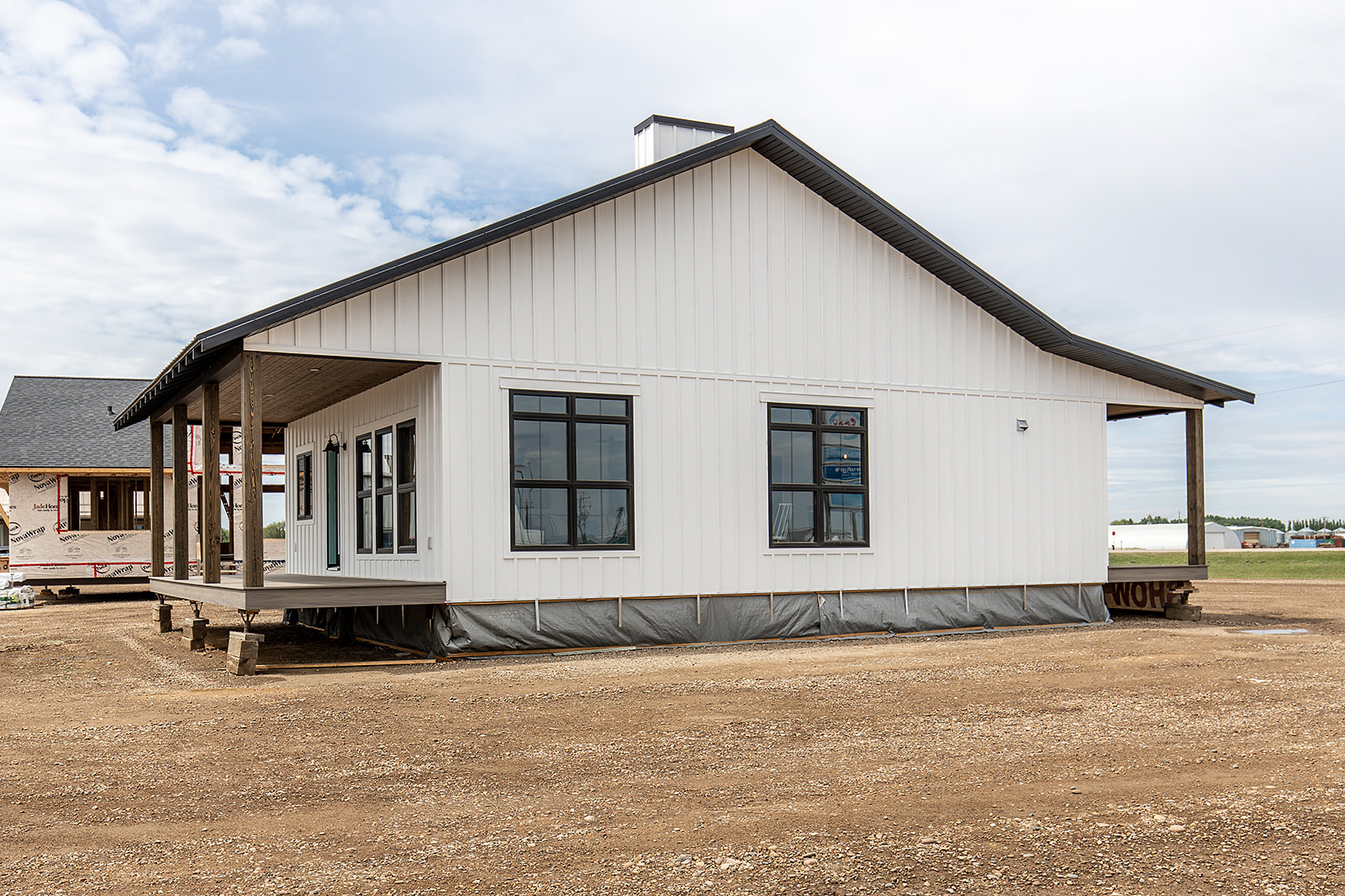 A white, modern prefab house with a sloped roof sits on a dirt lot. It has large black-framed windows and a covered porch, under a partly cloudy sky.