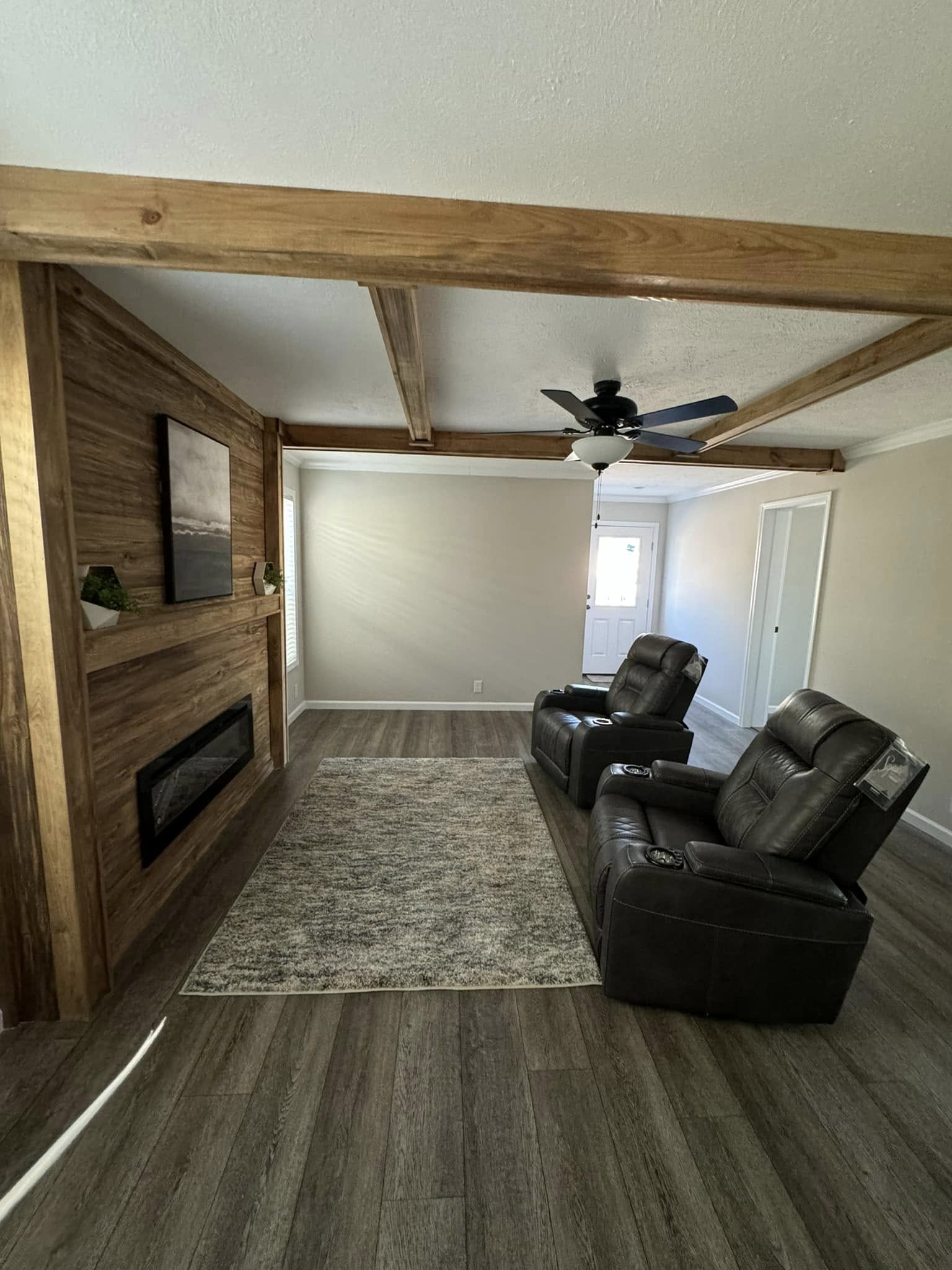 Modern living room with two brown recliners on a gray rug, wood-paneled wall with fireplace and art, exposed beams, ceiling fan, and soft natural light.