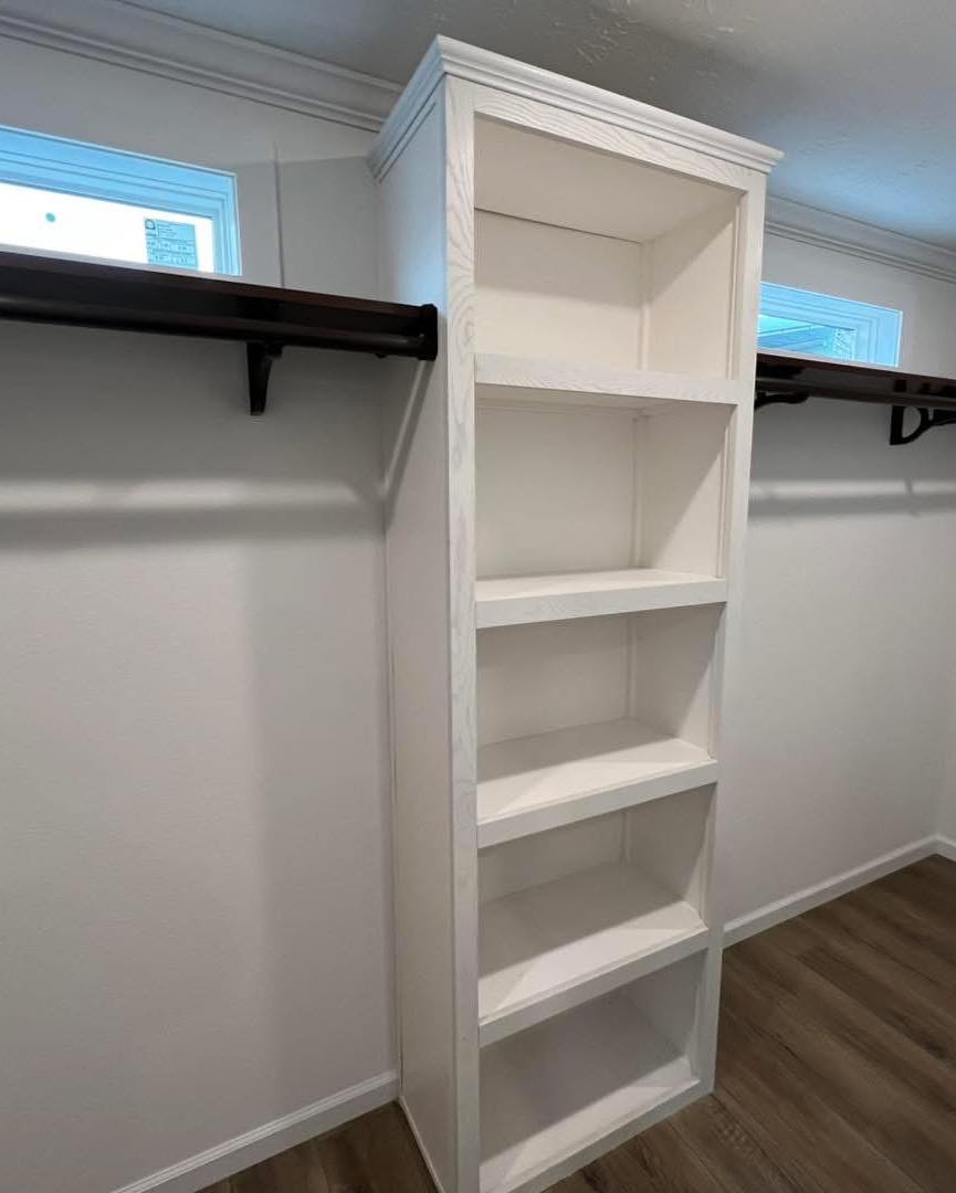 Tall white shelving unit in a closet with four shelves, surrounded by white walls and dark wooden clothing rods. Bright windows add natural light.