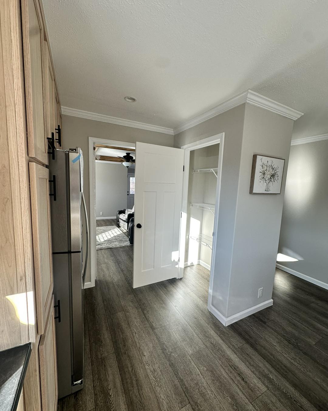 A modern kitchen hallway with light wood cabinets and stainless steel fridge on the left. A white door opens into a room with a ceiling fan and window.