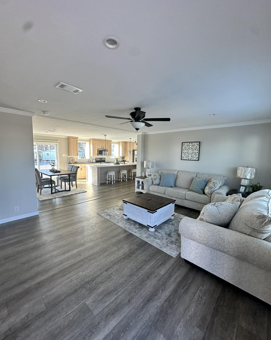Spacious living room with light gray walls, featuring a cozy beige sofa, blue cushions, ceiling fan, and a view into a bright kitchen with wooden cabinets.