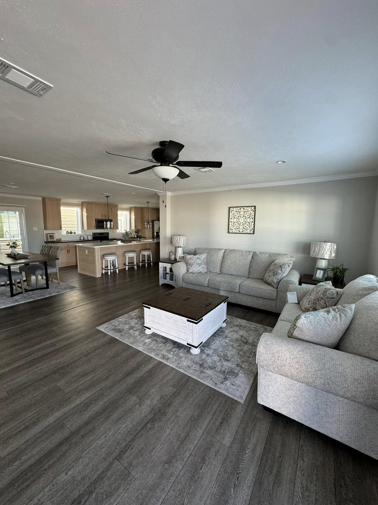 Spacious living area with gray sofas, a white coffee table, and dark wood flooring. Open kitchen with barstools and dining area in the background. Cozy ambiance.