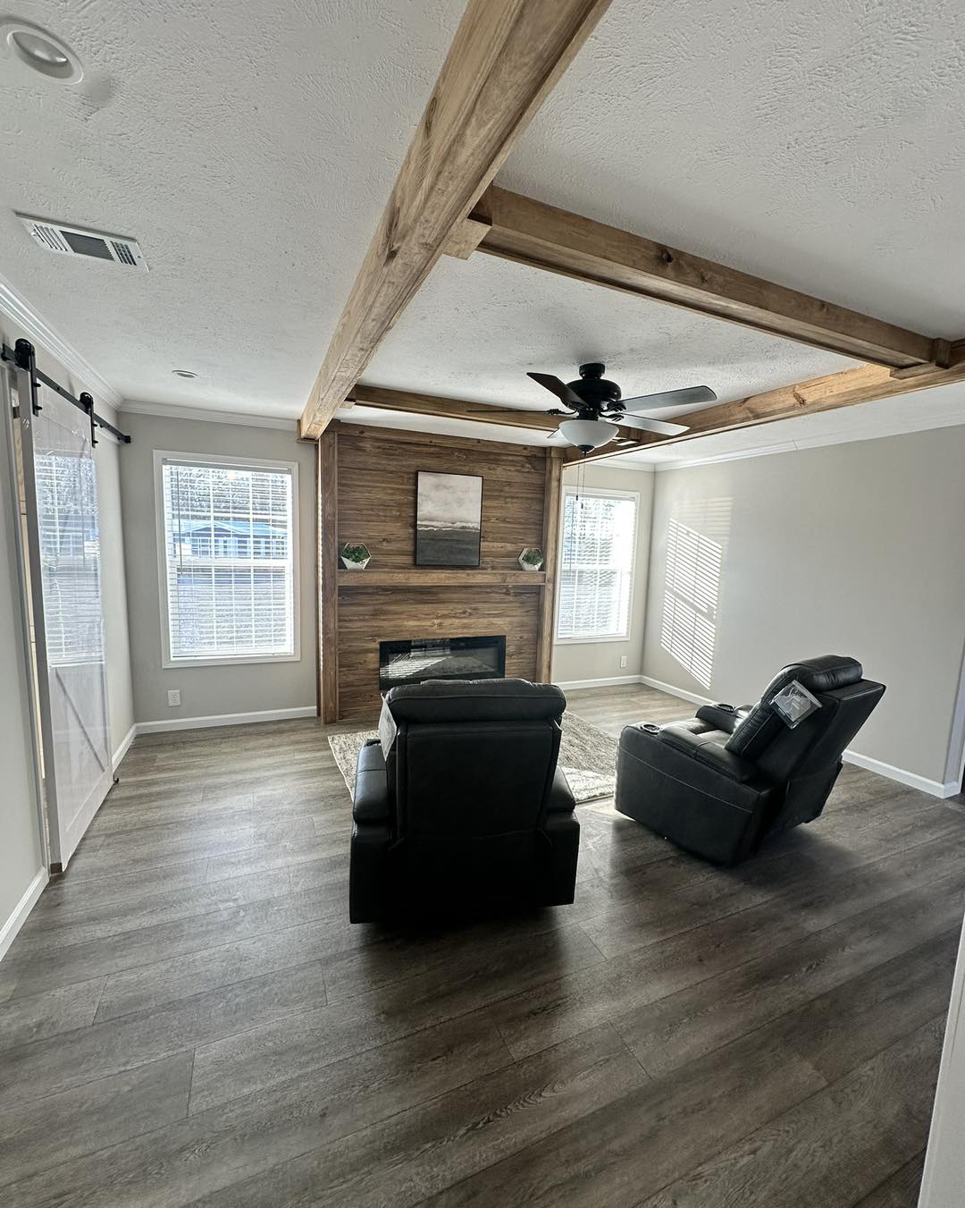 Modern living room with wooden beams on the ceiling, two plush chairs facing a wooden accent wall with a fireplace, and two large windows letting in natural light.