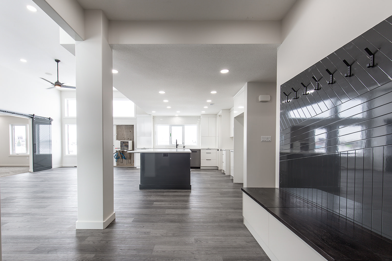 Spacious modern living area with gray hardwood floors, white walls, large windows, a black ceiling fan, and a sliding barn door, conveying minimalism.