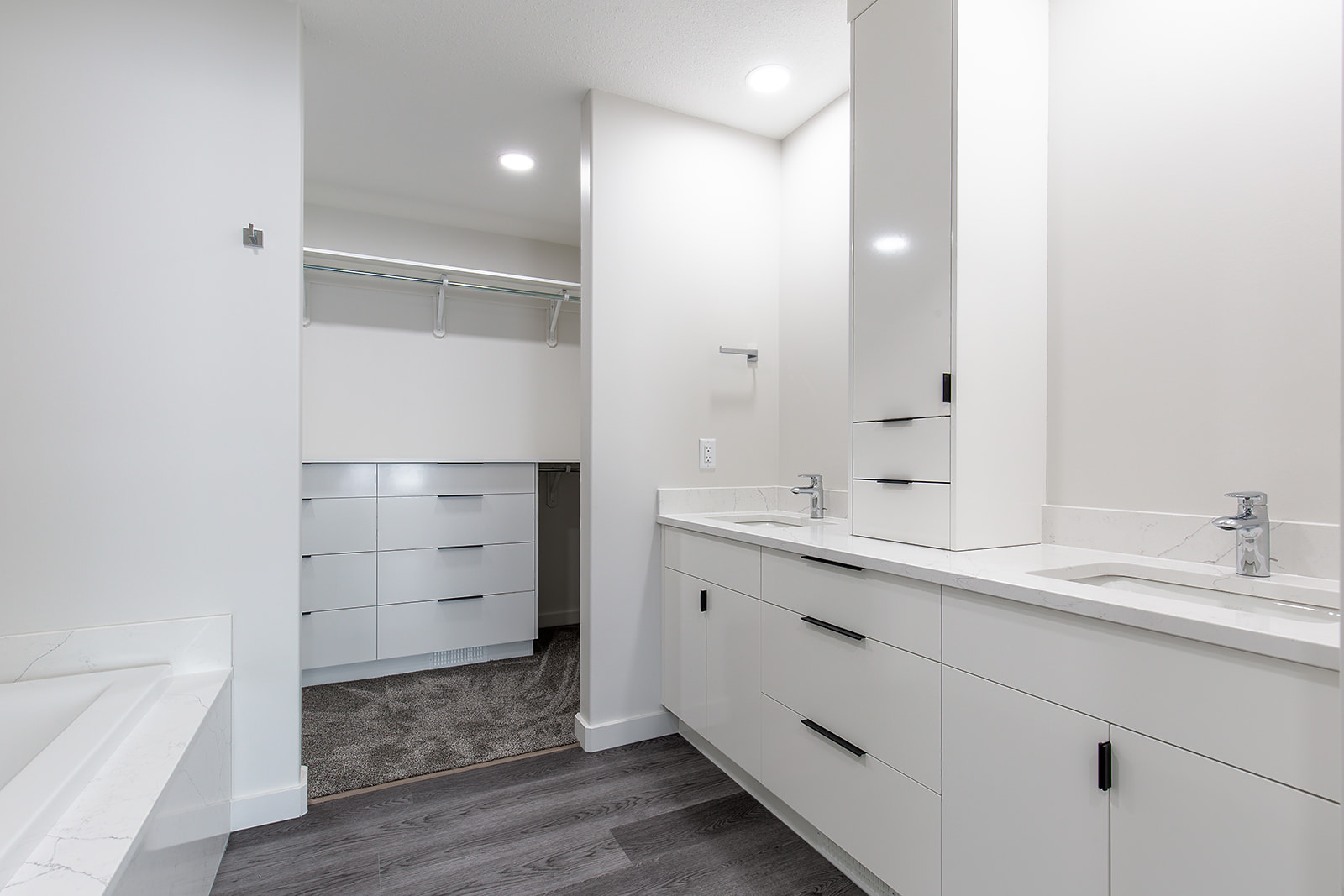 Spacious, modern foyer with white walls and grey wood flooring. Features a black front door, wall hooks above a bench, and a hallway leading to stairs.