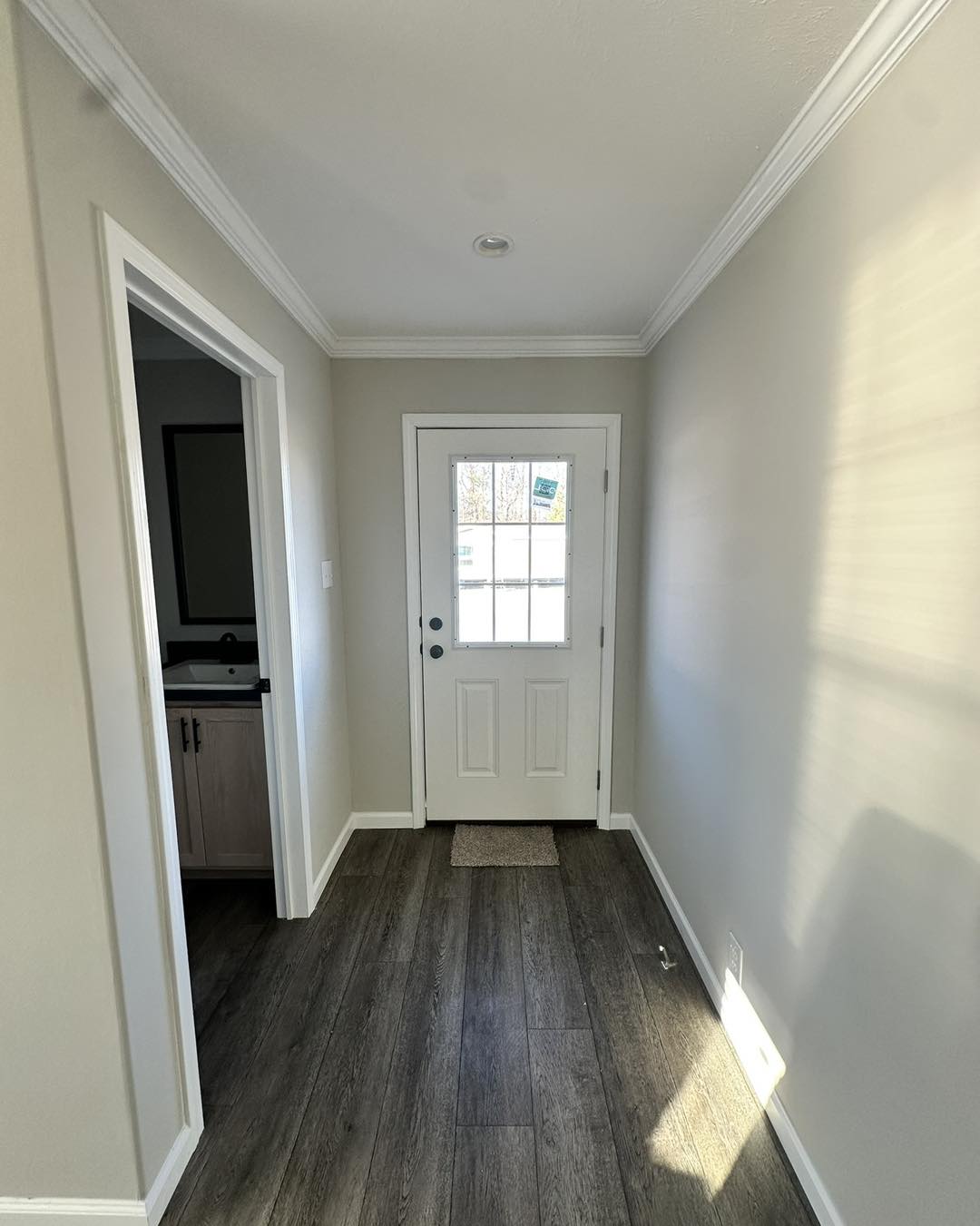 Narrow, bright hallway with light gray walls and wood flooring, leading to a white door with glass panes. A small bathroom is visible on the left.