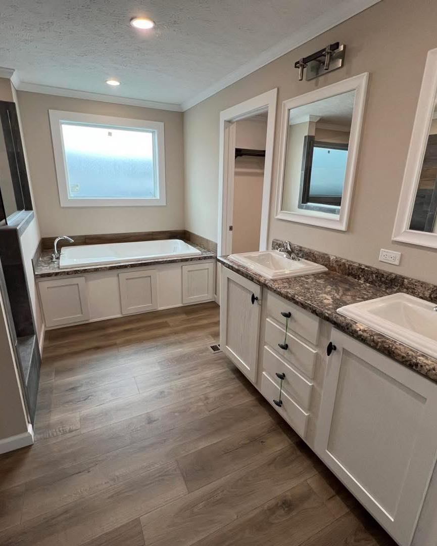 Modern bathroom with light wooden floor, featuring a bathtub under a large frosted window. Double sinks with marble countertops provide an elegant touch.