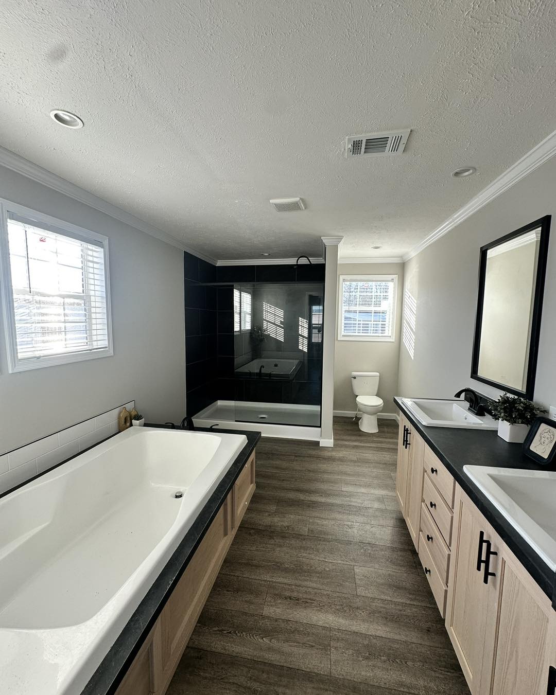 Modern bathroom with wood flooring, featuring a large bathtub, double sink vanity, shower, and toilet. Ample natural light from two windows creates an airy feel.