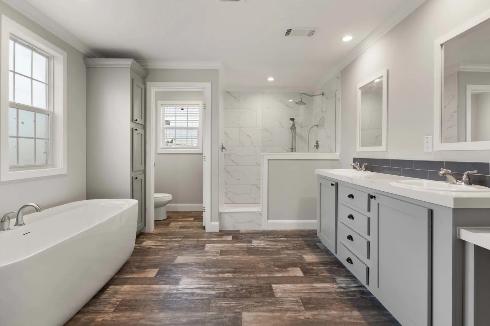 Spacious bathroom with light gray walls, a freestanding bathtub, wood-look tile flooring, dual sinks with mirrors, and a large shower area.