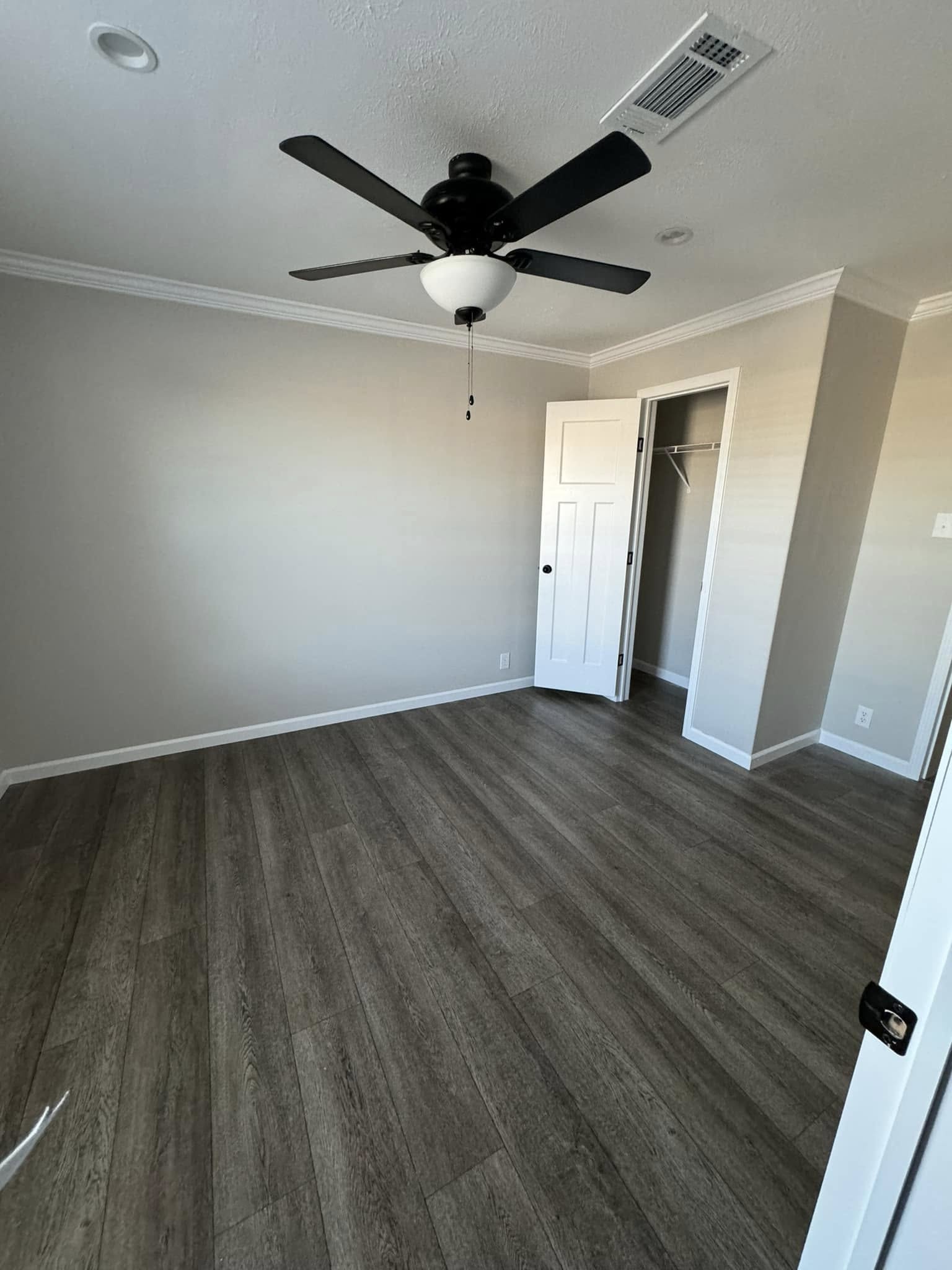 Empty room with gray walls and wooden floor, featuring a black ceiling fan. An open closet door reveals a small, bare closet space.