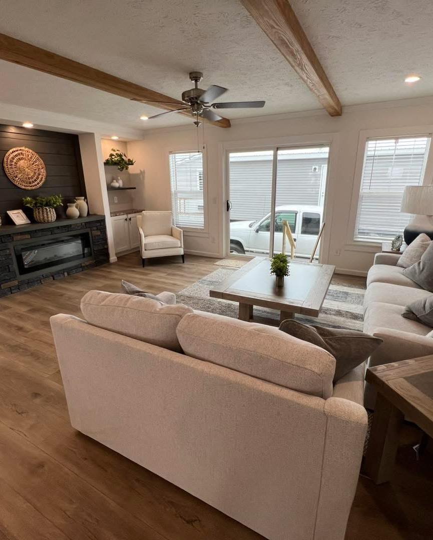 Cozy living room with light beige sofas, wooden coffee table, and soft lighting. A fireplace is on the left. Large windows provide natural light.