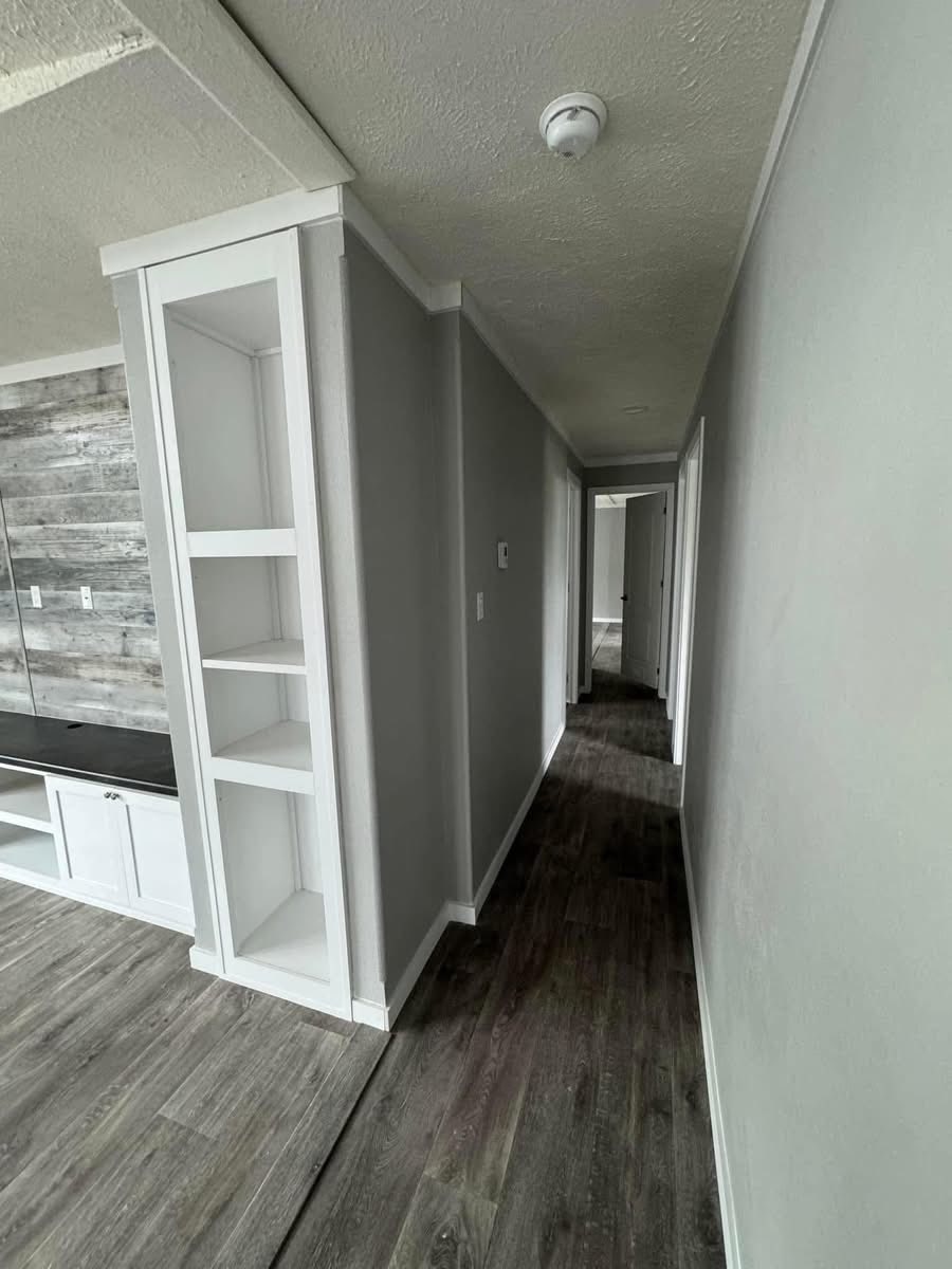 A narrow hallway with gray walls and wood flooring, leading to a distant door. The left side features open white shelves and a rustic wood accent wall.