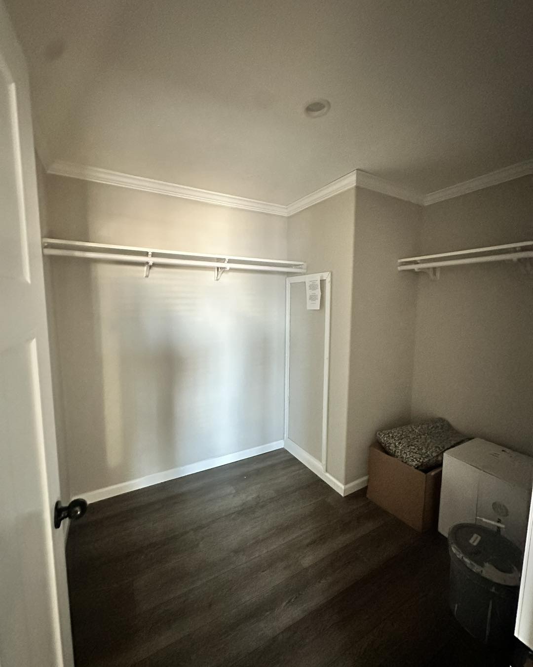 Empty closet with light gray walls, dark wooden floor, and white shelves on three sides. A brown box and a patterned item sit in the corner, next to a trash can.