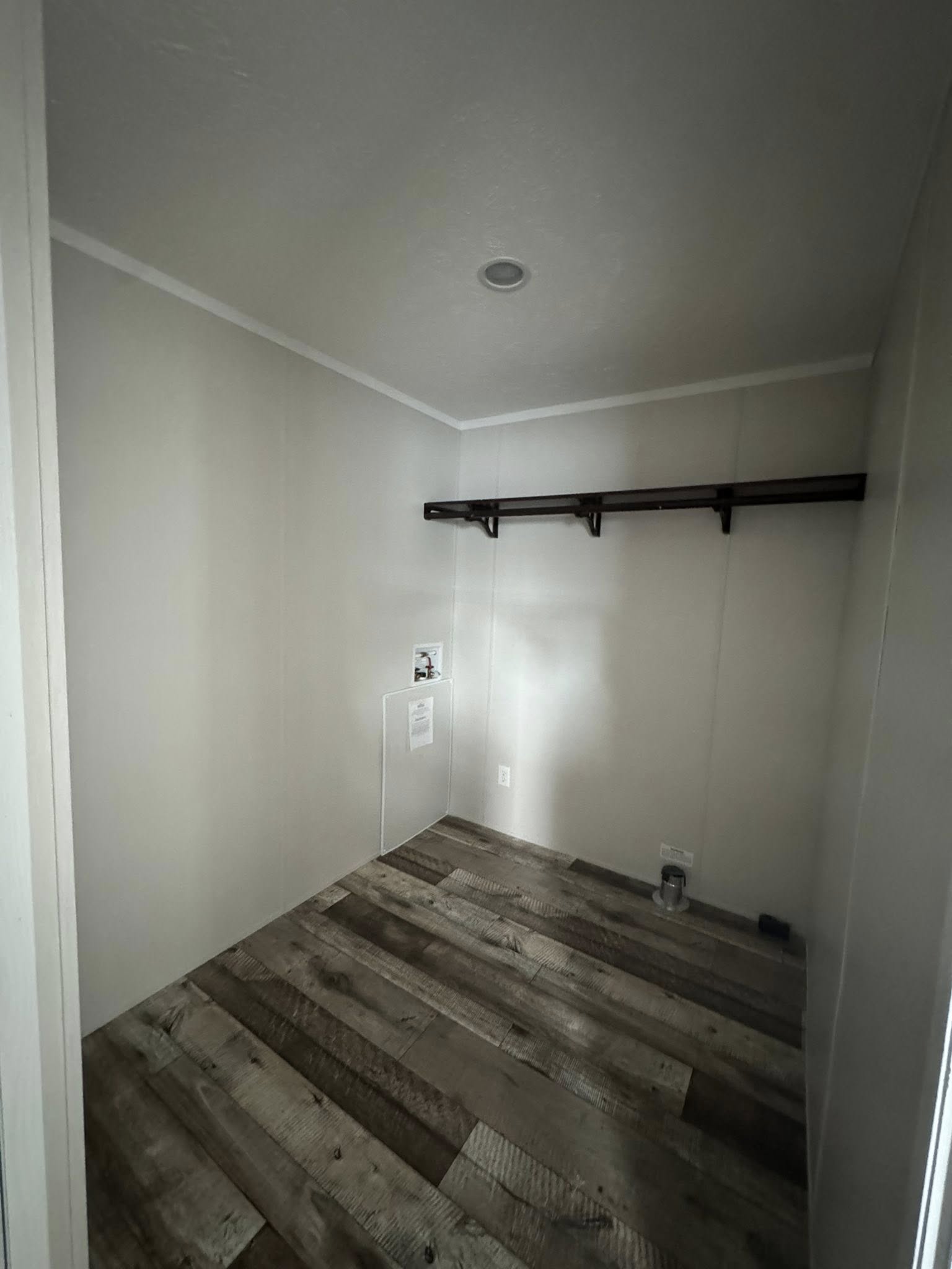 Empty room with wooden laminate flooring, light gray walls, and a narrow wooden shelf on one side. A small utility box is visible on the wall.