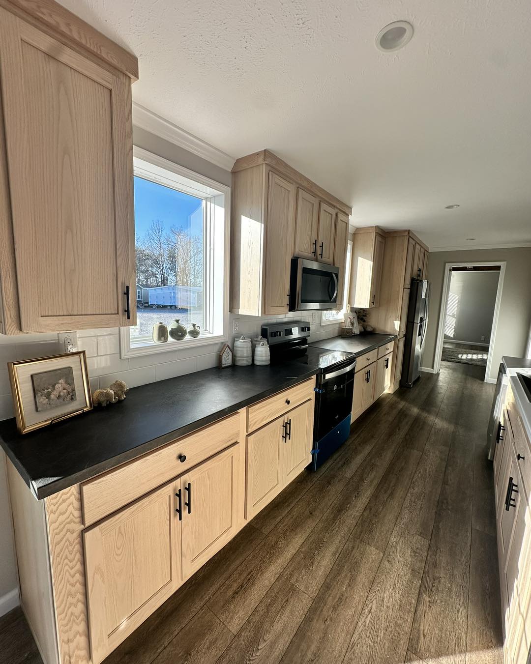 Bright kitchen with wooden cabinets, dark countertops, and hardwood floors. Sunlight streams through a window, creating a warm, inviting atmosphere.