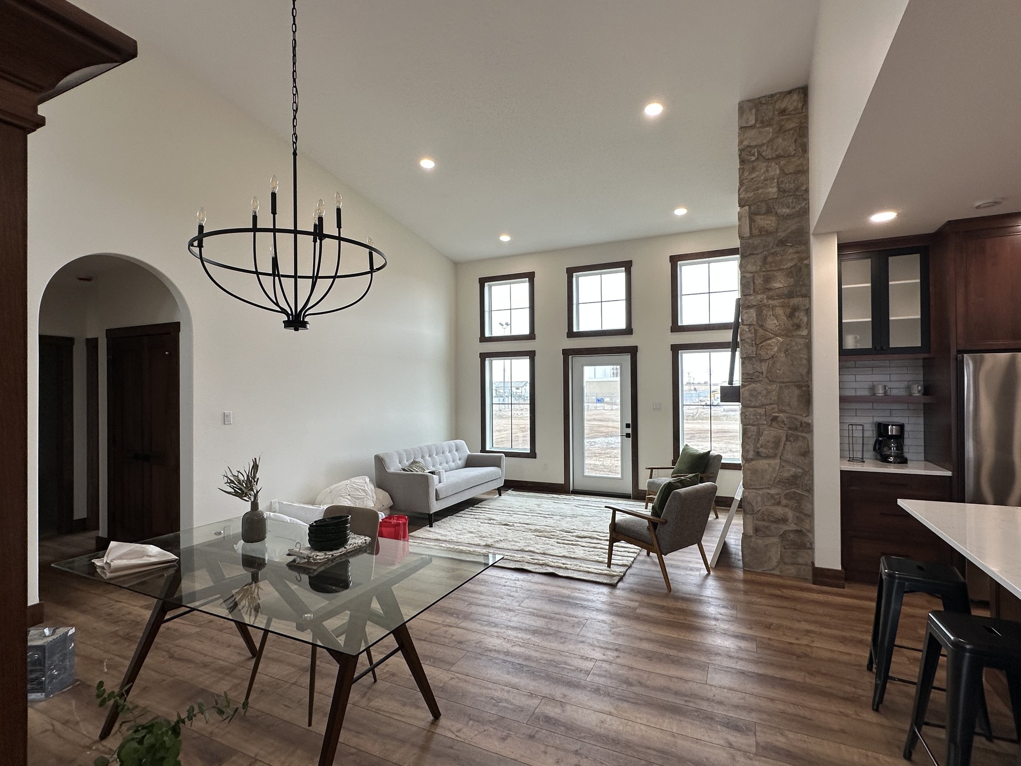 Modern living and dining area with large windows, wood flooring, and a stone accent wall. Features a gray couch, glass table, and minimalist decor. Bright and airy atmosphere.