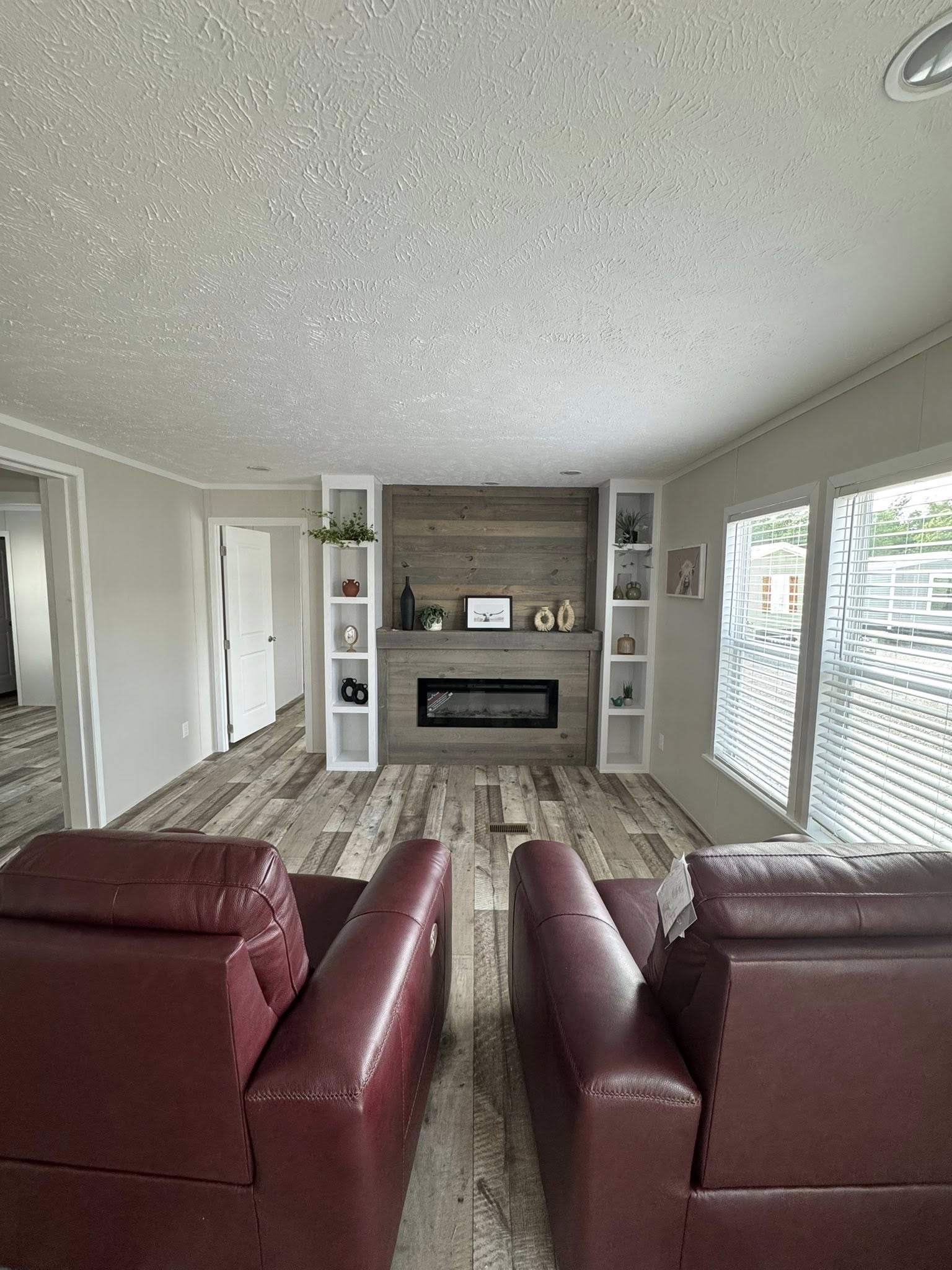 A cozy living room with two red armchairs facing a modern fireplace. Shelves with decor flank the fireplace. Large windows brighten the wood-accented space.