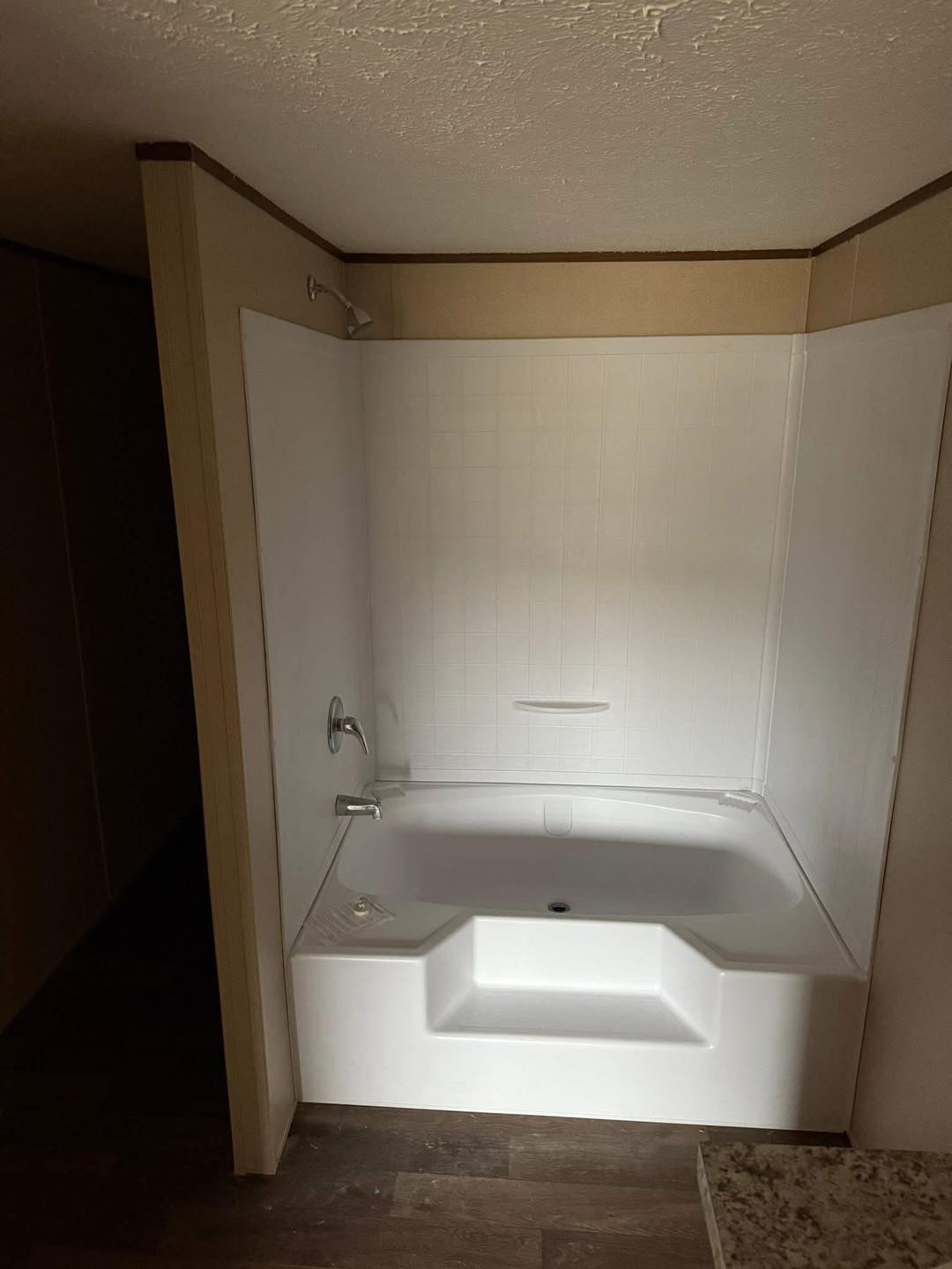 A white bathtub with built-in steps is set against a white-tiled wall in a dimly lit bathroom. The brown wooden floor and beige walls add warmth.
