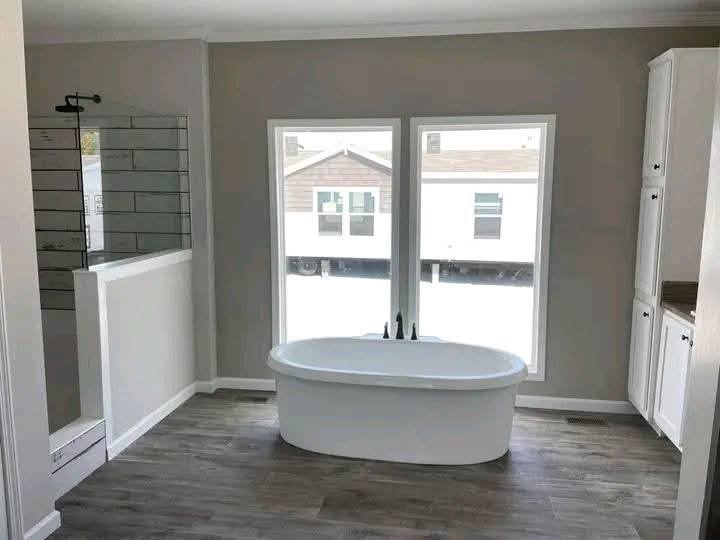 Modern bathroom with a central freestanding bathtub, large windows, and wood-like flooring. A glass-enclosed shower is on the left, creating a serene, airy feel.