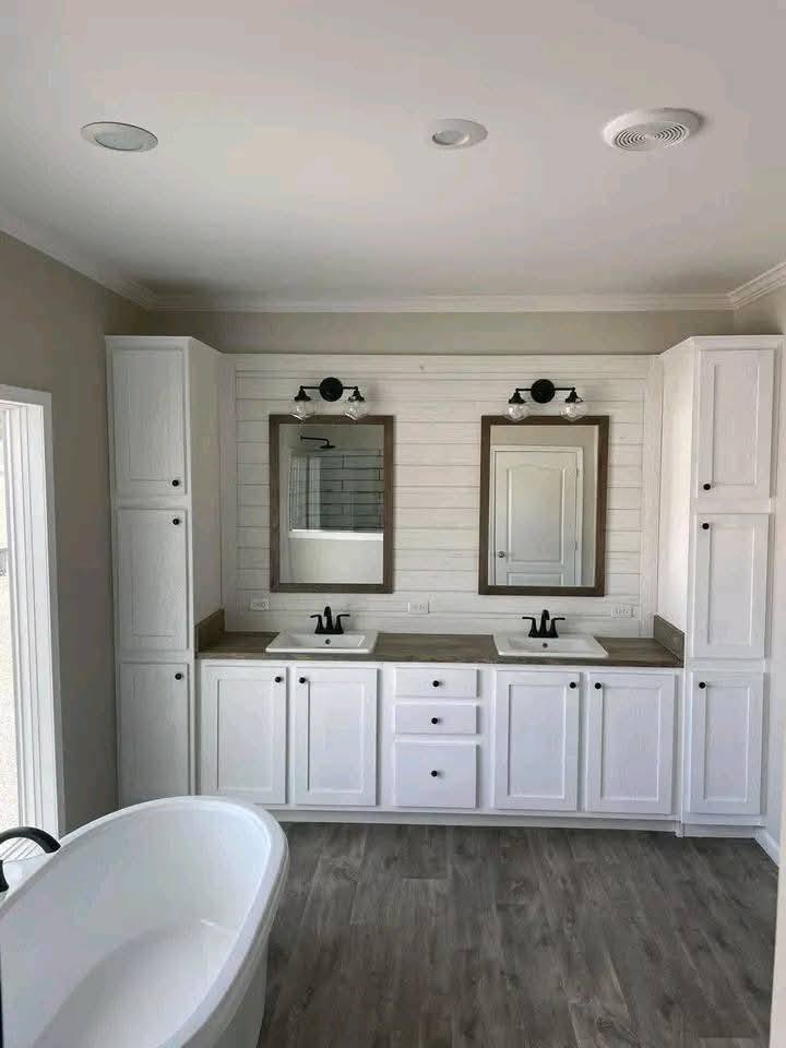 Spacious bathroom with dual sinks, dark faucets, and framed mirrors on shiplap wall. White cabinetry and a standalone bathtub on wooden flooring.
