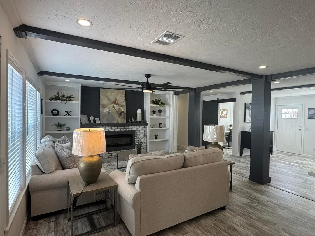 Modern living room with beige sofas facing a brick fireplace. Shelves with decor surround the fireplace. A ceiling fan and large windows add a spacious feel.