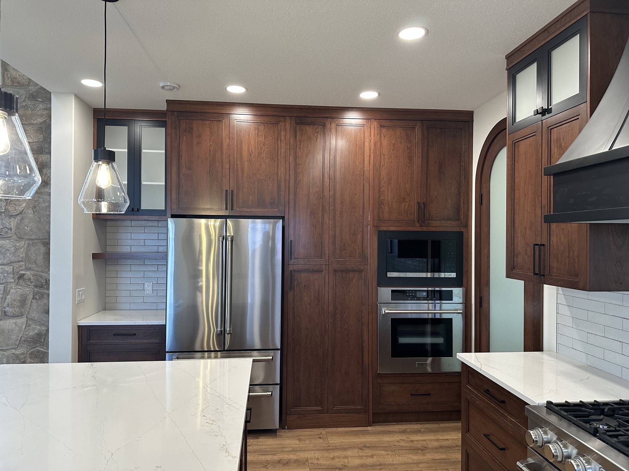 Modern kitchen with dark wood cabinets, stainless steel refrigerator, double oven, and white marble countertops. Warm lighting and stone accents set a cozy tone.