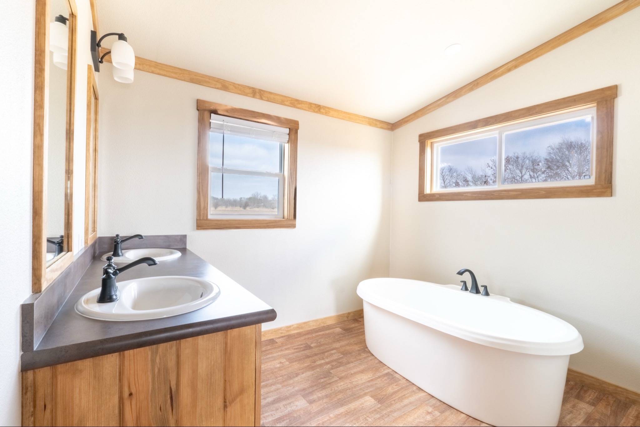 Bright, airy bathroom with wooden accents. Features a white freestanding tub, dual sinks with black faucets on a wooden vanity, and large windows.