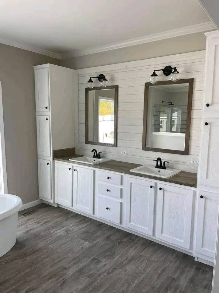 Spacious bathroom with dual sinks, black faucets, and white shaker cabinets. Above are two framed mirrors and industrial light fixtures. Wood flooring.