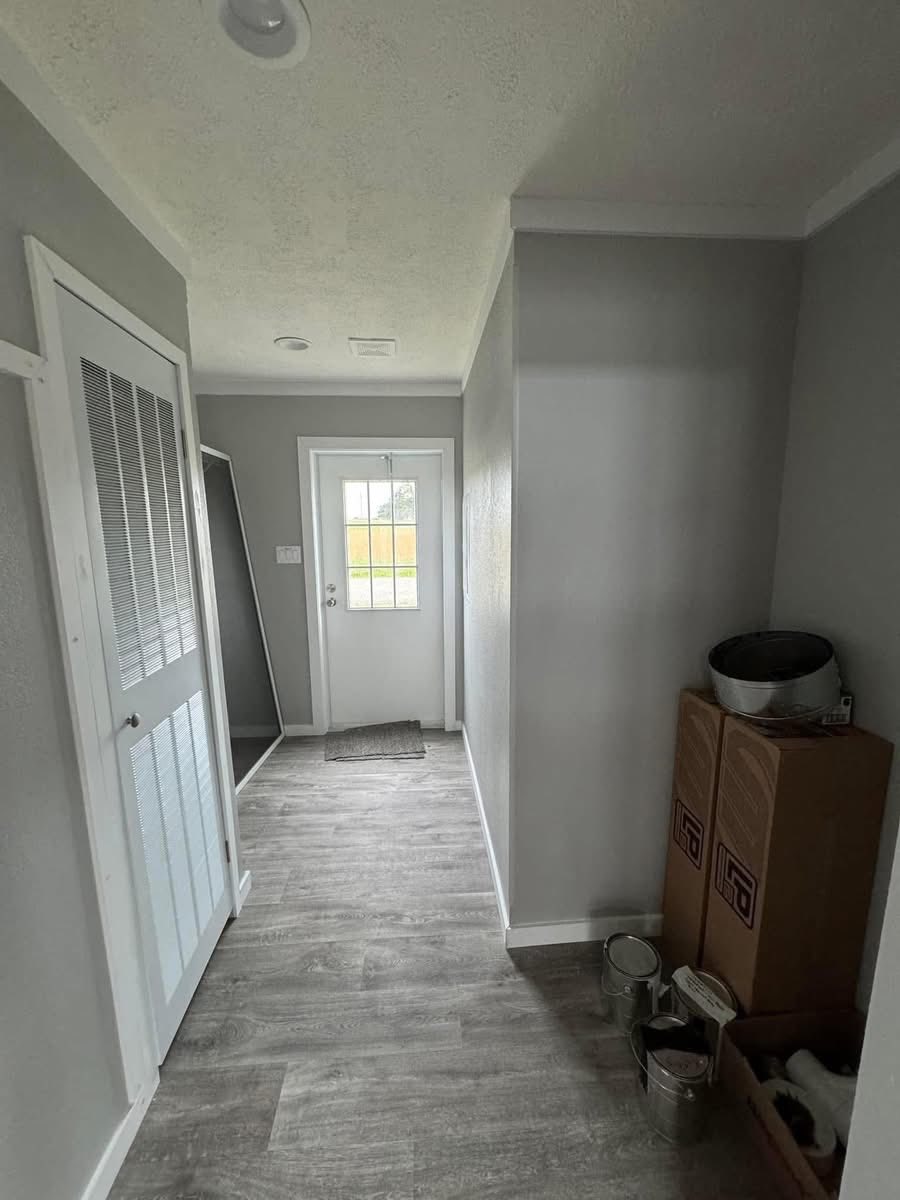A narrow, gray-painted hallway with wooden flooring leads to a glass door. On the right, boxes and paint cans are stacked, suggesting a recent renovation.
