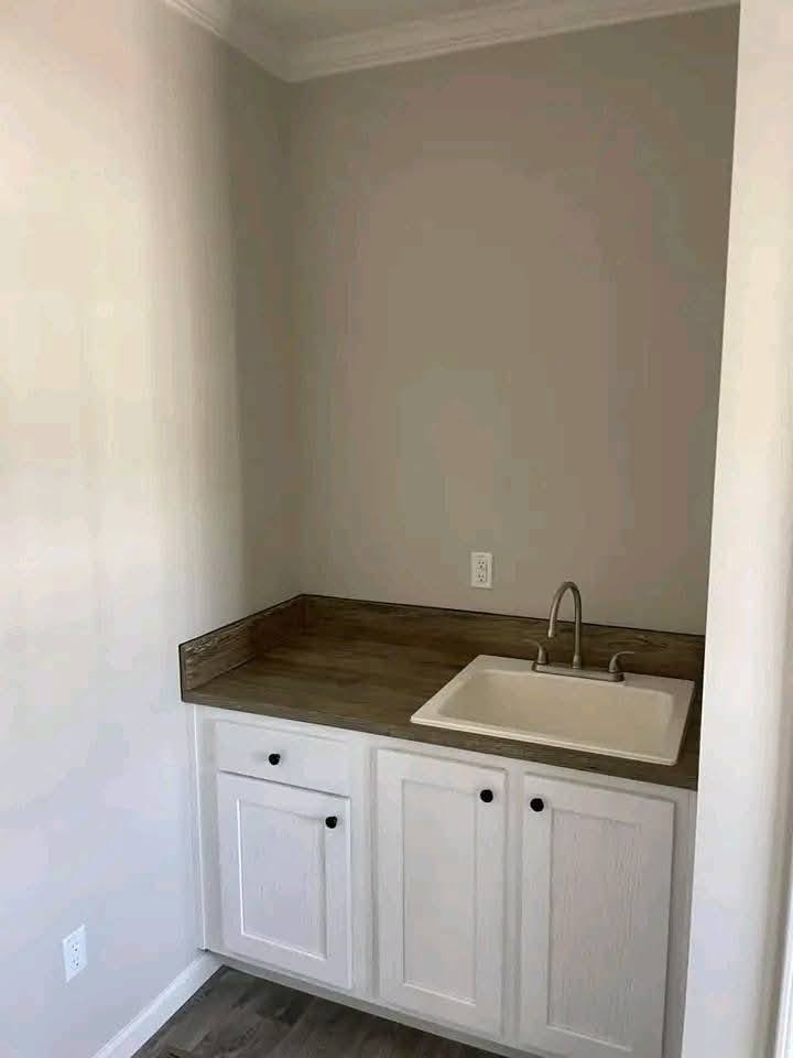 A small laundry area with neutral tones features a wooden countertop, square white sink, and white cabinets. The space conveys simplicity and functionality.