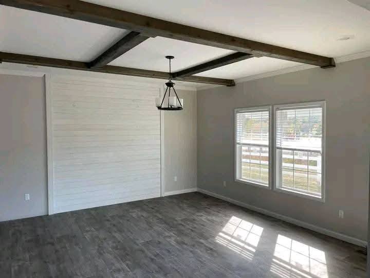 A spacious, empty room with light gray walls, wooden beams on the ceiling, a black metal chandelier, and two double windows letting in natural light.
