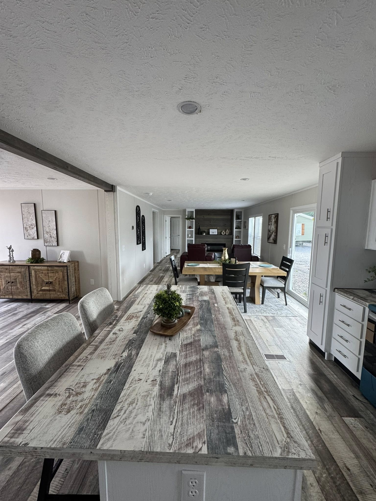 Long, rustic-style dining table with a plant centerpiece, leading to an open living area. Cozy chairs, wall art, and large windows create an inviting space.
