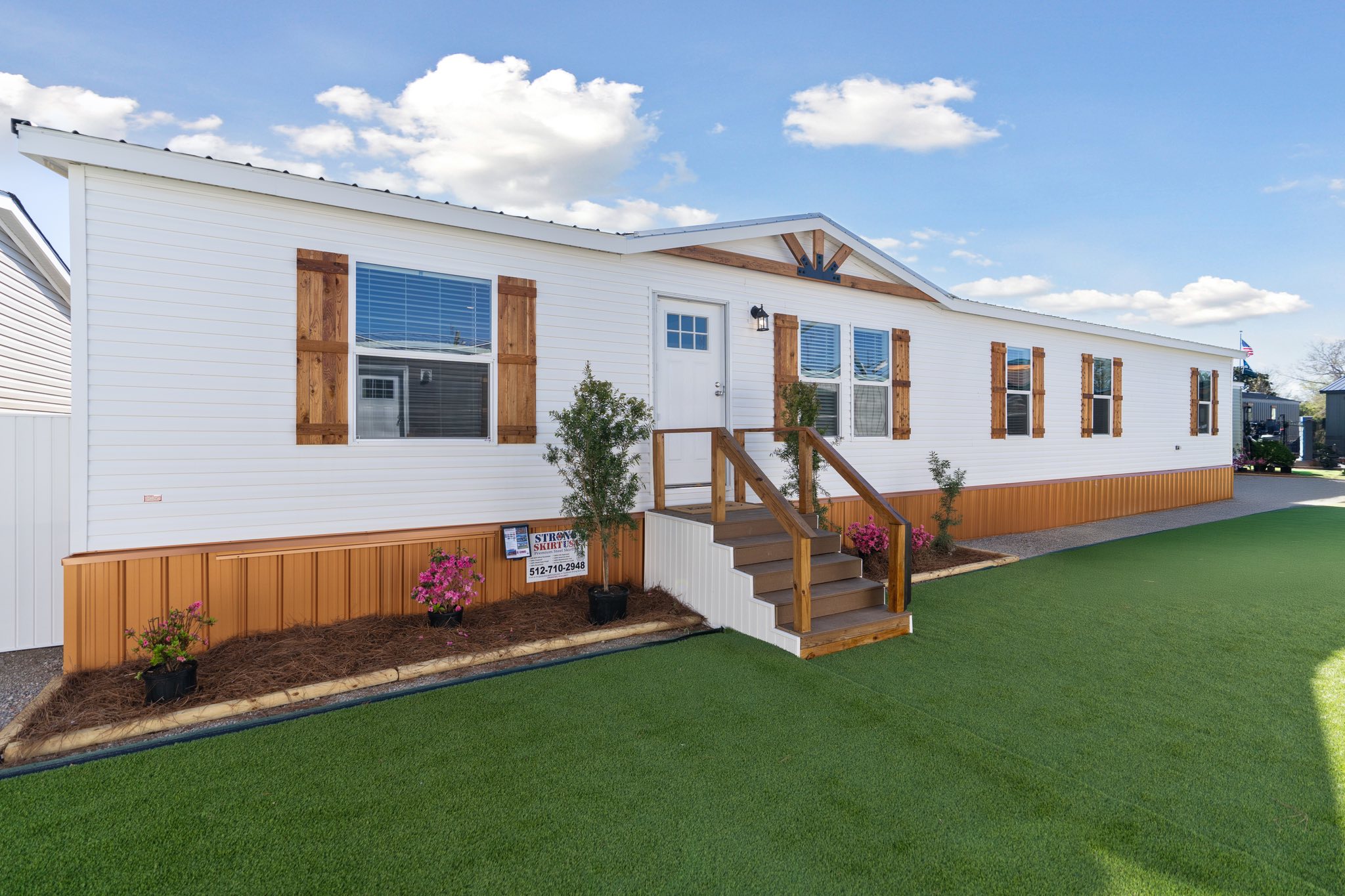 A white mobile home with wooden shutters and a covered entrance, set against a clear blue sky. Neat landscaping includes flowers and artificial grass.